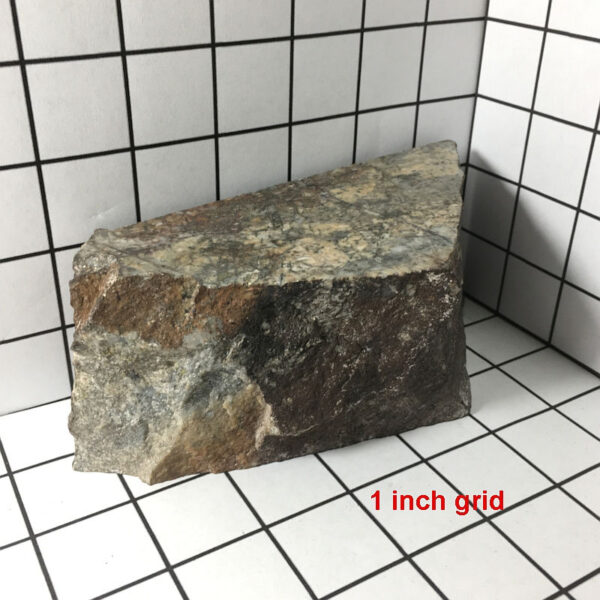 A piece of Breccia de Oro rough rock with three cut faces on a one inch grid. Approximate measurements are 6.5 by 2.5 by 2 inches.